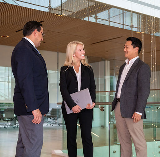 A smiling woman with a laptop stands between two men in an office.