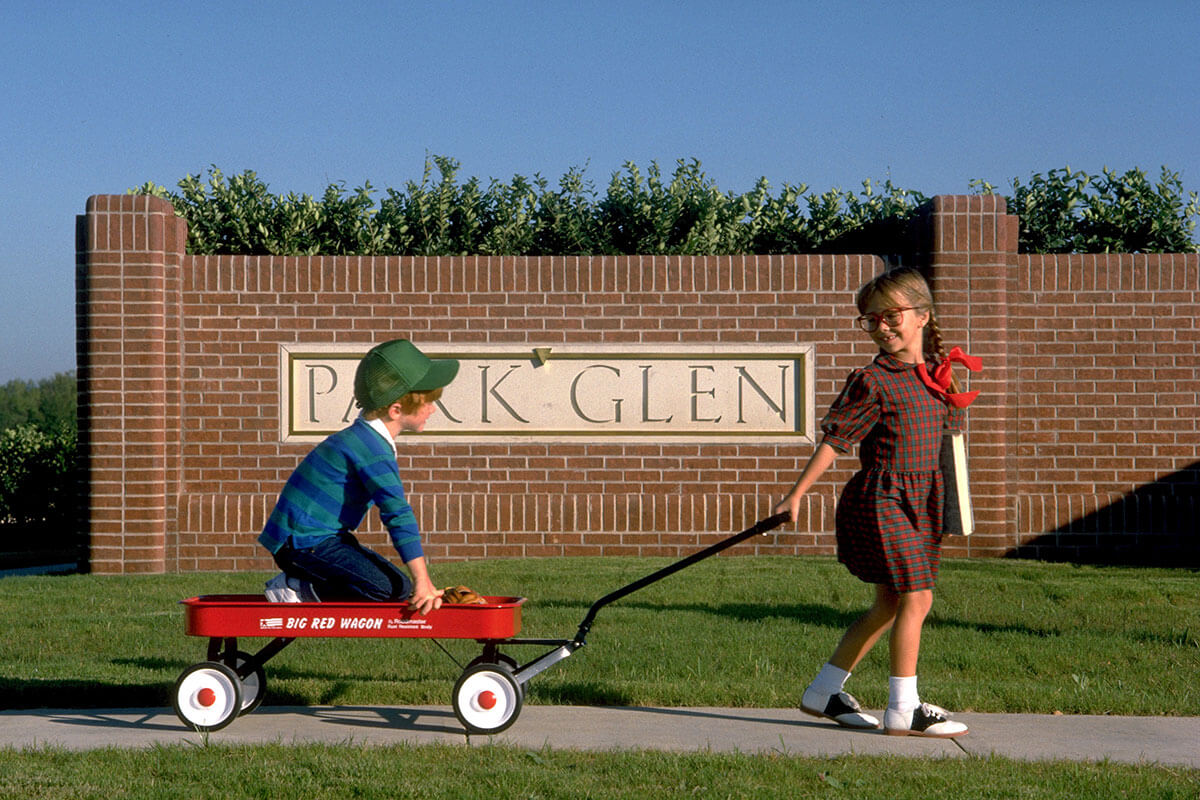 A young girl pulls a red wagon holding a young boy in front of a brick sign reading, 'Park Glen'.