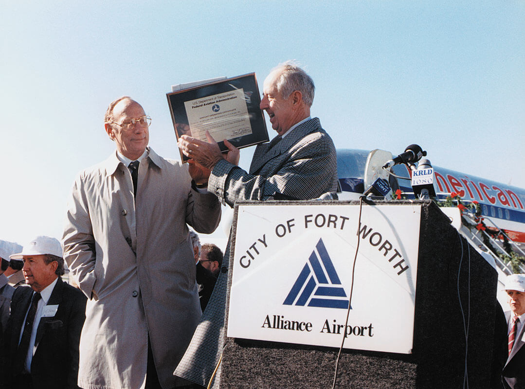 Two men speak at an outdoor lectern that displays a sign reading, 'City of Fort Worth Alliance Airport'.