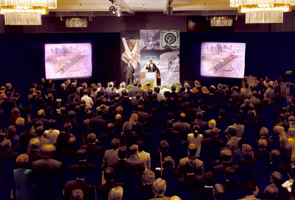 A crowd of people is seen from behind as they watch a presentation on two large screens.