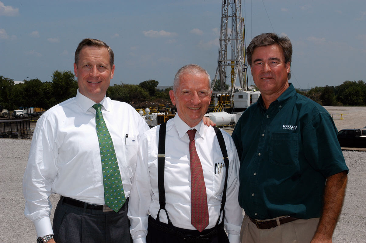 Ross Perot Jr., Ross Perot Sr. and a representative of Chief Oil & Gas are seen in front of an oil rig.