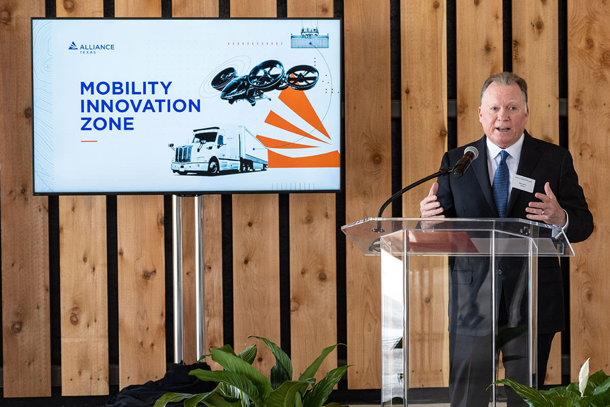 A man at a lectern speaks beside a screen reading, 'Mobility Innovation Zone'